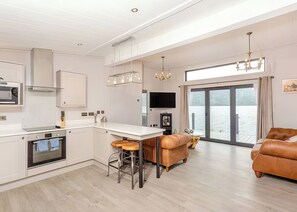 A modern kitchen with a large island, a dining area with a sofa, and a view of the water through large windows. - 1 bedroom accommodation in Invermoriston (Invermoriston)
