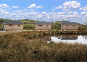 Three wooden cabins with sloped roofs, situated by a calm pond with tall grasses, under a partly cloudy sky. - The studio lodge is perfect for couples. (Ringford)