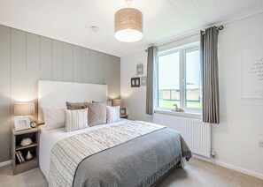 A bedroom with a bed, bedside lamps, a window with curtains, and a wall-mounted light fixture. - The Glades Lodges with Hot Tubs (Pet) (Westbury)