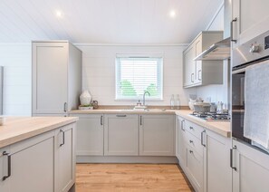 A modern kitchen with light grey cabinets, a wooden countertop, a stainless steel refrigerator, and a window with blinds. - The Glades Lodges with Hot Tubs (Pet) (Westbury)