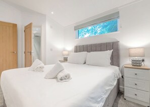 Ein ordentlich bezogenes Bett mit weißen Leinen und Handtüchern, ein grauer möbelbezogener Kopfteil, eine Nachttischlampe und ein Nachttisch mit Schubladen.
