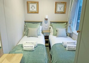A twin room with two beds, each with a pillow and a towel, a bedside table, a nightstand, and framed artwork on the wall. - Premium 2 bedroom holiday home with hot tub (Bassenthwaite, near Keswick)