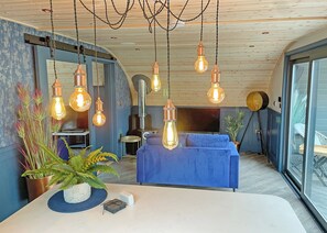 A modern living room with a blue sofa, a dining table with a plant, and multiple hanging light fixtures. - Glamping Pod with hot tub - Pet Friendly (Bassenthwaite, near Keswick)