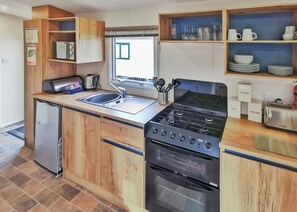 A compact kitchen with a built-in stove, oven, and sink. There are wooden cabinets, a small refrigerator, and a window with a view. - Gold Plus 3 No Dogs Sleeps 6 (Fishguard, Pembrokeshire)