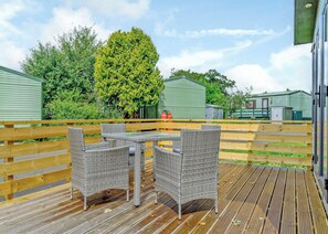Eine Holzterrasse mit Korbmöbeln und einem Tisch, umgeben von grünen Fertighäusern.