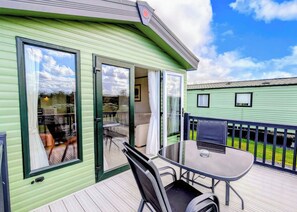 A green mobile home with a patio area featuring a table and chairs, a sliding glass door, and a small balcony. - Tweed Spa + - 2 Bedroom (Pet Friendly) (Coldstream)