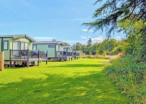 A row of green mobile homes with wooden decks and windows, set in a grassy area with trees in the background. - Tweed Spa + - 3 Bedroom (Pet Friendly) (Coldstream)