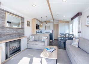 A modern living room with a fireplace, sofa, and coffee table, connected to a kitchen and dining area with wooden accents and a large mirror. - Spa Luxe - 3 Bedroom (Coldstream)