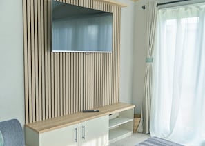 Ein modernes Wohnzimmer mit einem an einer Holzvertäfelung befestigten Flachbildfernseher, einer hellen Unterhaltungseinrichtung mit Fernbedienung und einem großen Fenster mit transparenten Vorhängen.