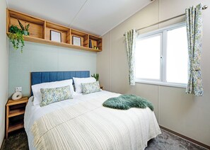 A bedroom with a bed, a nightstand, a wooden shelf with framed pictures, a window with curtains, and a wall-mounted plant. - Luxury 2 Bedroom Holiday Home – Pet Friendly (Bassenthwaite, near Keswick)