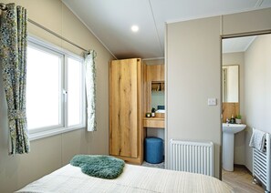A bedroom with a bed, a wooden wardrobe, a window with curtains, a radiator, and a bathroom with a sink and mirror. - Luxury 2 Bedroom Holiday Home – Pet Friendly (Bassenthwaite, near Keswick)