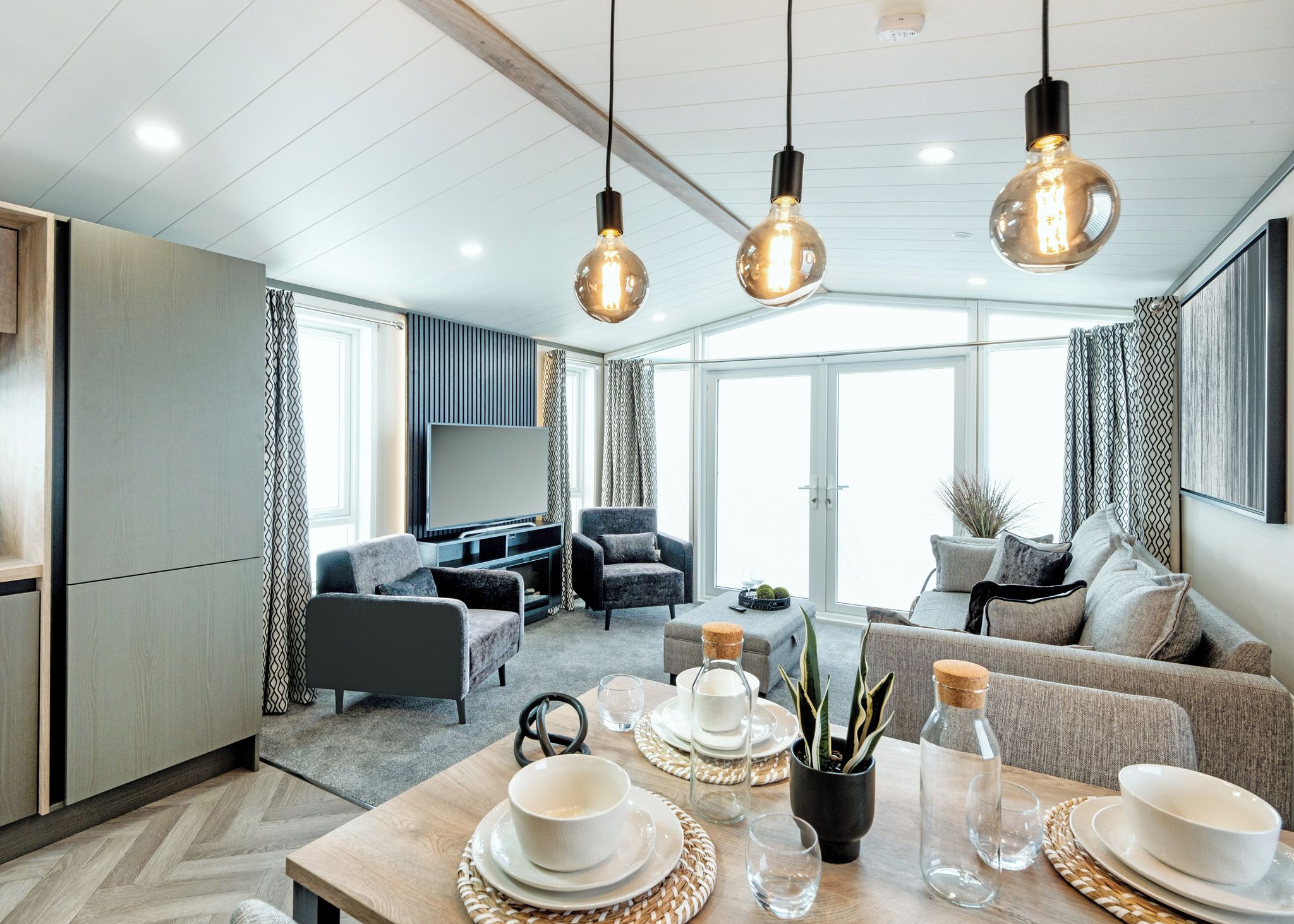 Een moderne woonkamer met een bank, fauteuils, een eettafel gedekt met borden en glazen, en hanglampen.