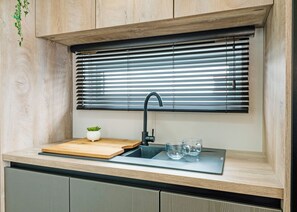 A kitchen with a built-in sink, a wooden cutting board, a potted plant, and a window with horizontal blinds. - Premium 2 Bedroom Holiday Home - Pet Friendly (Bassenthwaite, near Keswick)