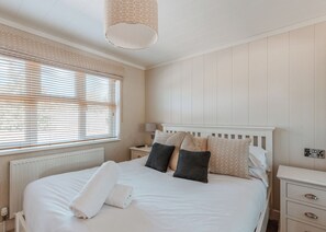 A neatly made bed with pillows and a rolled towel, a bedside table with a lamp, and a window with blinds. - Cherry Lodge VIP Hot Tub and Pet Friendly (Bridgnorth)