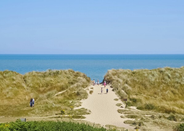 Ein sandiger Pfad führt zum Meer, vorbei an grasbewachsenen Dünen, während Menschen darauf spazieren.