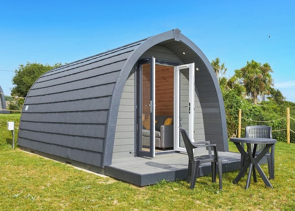 A small, single-story, semi-circular structure with a door leading to an interior space, a small patio with a table and chairs, and a grassy area with trees in the background. - 0 bedroom accommodation in Hemsby (Hemsby)