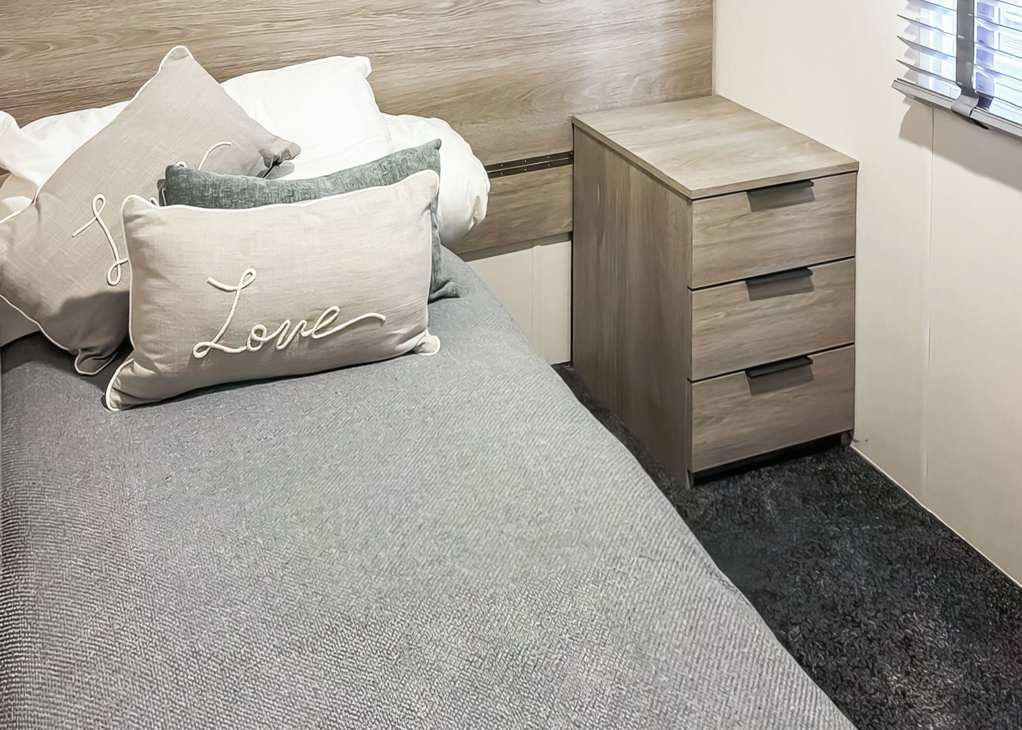 A neatly made bed with a grey mattress and pillows, a wooden bedside table with drawers, and a window with blinds.