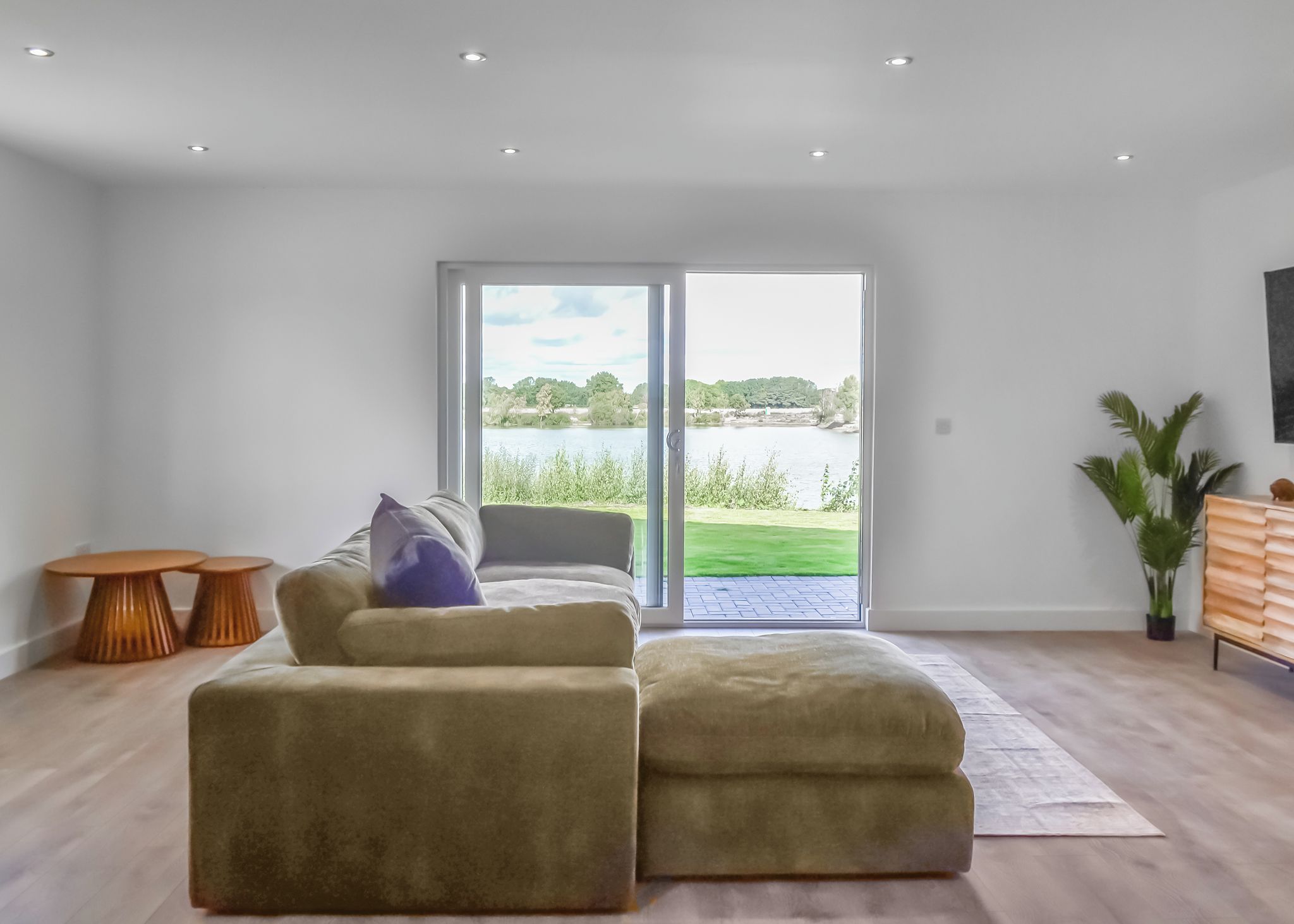 A modern living room with a sofa, a coffee table, and a television. There is a sliding glass door leading to a balcony with a view of a lake and greenery.