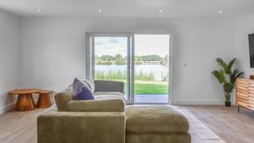 A modern living room with a sofa, a coffee table, and a television. There is a sliding glass door leading to a balcony with a view of a lake and greenery.