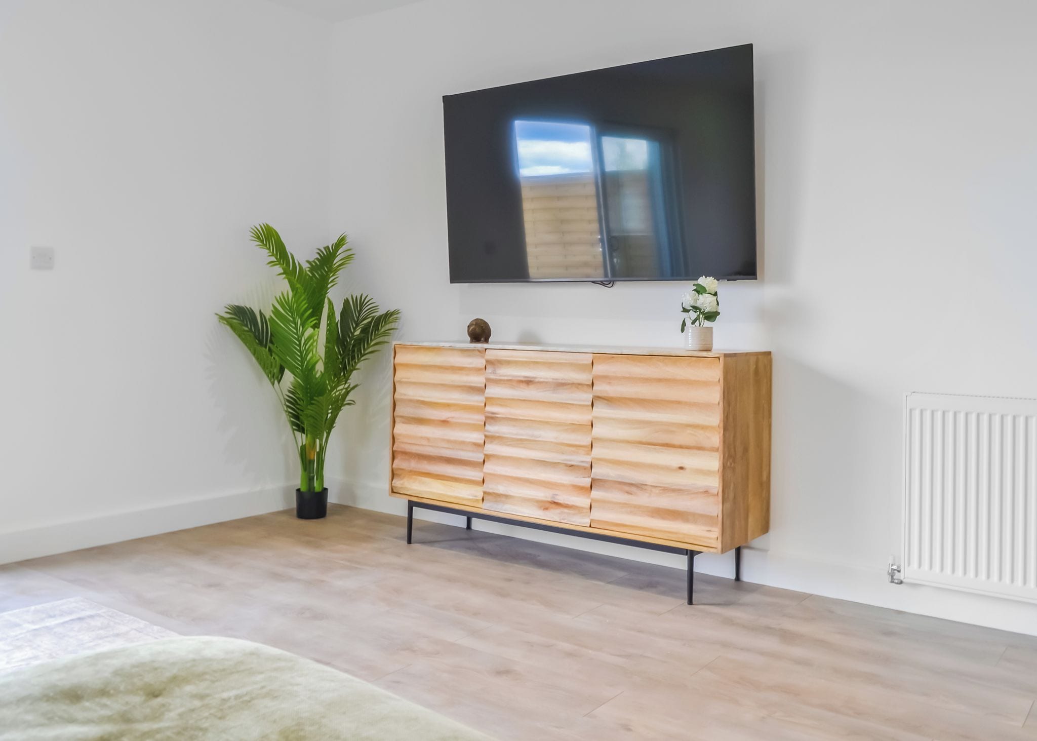 A modern living room with a wooden cabinet, a flat-screen TV mounted on the wall, and a potted plant.