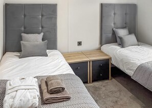 A hotel room with two beds, each with a grey headboard and white bedding. There is a wooden nightstand with drawers between the beds. - Signature Plus2 bedroom lodge hot tub pet friendly (Anstruther)