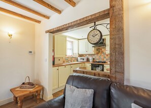 A living room with a leather sofa, wooden beams, a wall-mounted clock, and a view into a kitchen with a tiled backsplash. - The Dairy is a charming one-bedroom barn conversion at High Farm Holiday Park. (Beverley)
