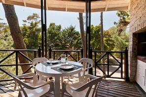 Un espace de restauration en plein air avec une table dressée pour deux personnes, entourée de chaises blanches, sous une pergola en bois offrant une vue sur des arbres et un bâtiment en pierre.