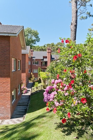 A brick building with a staircase leading to a second floor, surrounded by green grass and flowering bushes. - Las Cuatro Estaciones (Carilo)