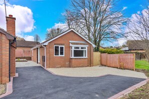 A single-story brick house with a front porch, a small attached garage, and a gravel driveway. - Finest Retreats | Charnwood Forest Lodge (Shepshed)