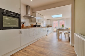 A modern kitchen with a built-in oven, microwave, and dishwasher. There is a dining area with a table and chairs, and a view of the outside through a large window. - Finest Retreats | Charnwood Forest Lodge (Shepshed)
