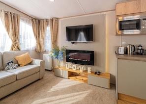 A modern living room with a flat-screen TV, a sofa, a coffee table, and a kitchen area with a microwave. - The Juniper Platinum is our signature caravan collection. (Dawlish)