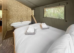 A bedroom with a large bed, two neatly folded towels, a bathtub, and a window with a view of greenery. - Hilltop Safari Lodge (Halling)