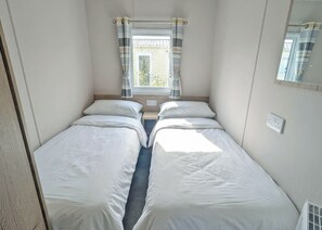 A compact room with two single beds, a window with curtains, a small bedside table, and a wall-mounted mirror. - Platinum 3 bed + Deck (Longridge, Ribble Valley)