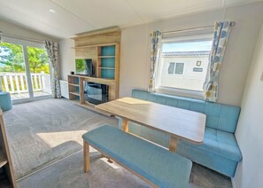 L’intérieur d’une caravane est aménagé avec une table en bois, des sièges bleus, un téléviseur et une fenêtre avec des rideaux.