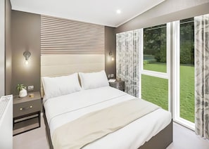 A modern bedroom with a large bed, bedside tables, a nightstand, and a view of a green garden through large windows. - Autograph S Pod Escape 4 Pet Friendly (Hot tub) (Thirsk)