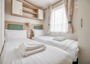 A hotel room with two beds, a wooden headboard, a wall-mounted shelf, and a window with floral curtains. - Kingfisher 2 Bed Hot tub (Pet) (Nr St Austell)