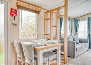 A dining area with a table set for four, two grey chairs, a fire blanket on the wall, and a window with a view of trees. - 1 bedroom accommodation in Nr St Austell (Nr St Austell)