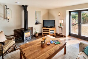 A cozy living room with a wood-burning stove, a television, and a coffee table with books and a candle. - Finest Retreats | Styles Farm Stables (Mappowder)