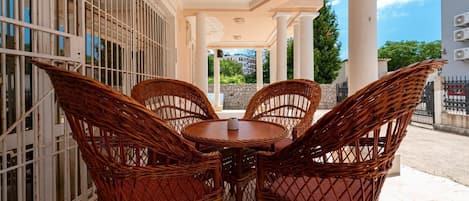 A covered outdoor seating area with wicker chairs and a table, overlooking a stone wall and greenery.