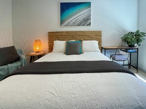 A neatly made bed with a patterned bedspread, a bedside lamp, a chair, a desk with a chair, and a potted plant.