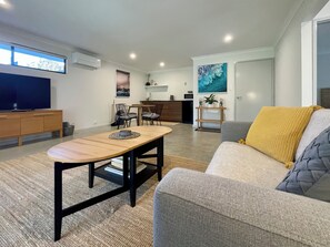 Ein modernes Wohnzimmer mit einer Couch, einem Couchtisch und einem Fernseher. Im Hintergrund ist eine Küchenzeile mit einem Esstisch und Stühlen zu sehen.