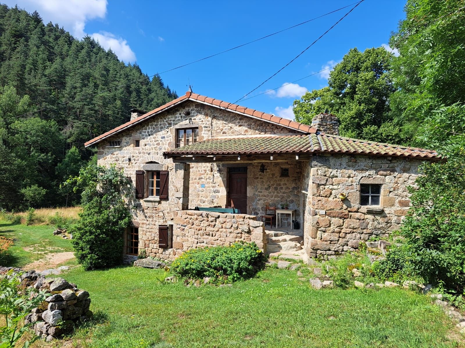 Une maison en pierre avec un toit de tuiles, entourée de verdure et adossée à une montagne.