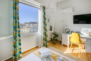 A hotel room with a bed, a desk, a chair, a TV, and a view of a cityscape. - SMG Boutique Apartment (Athina)