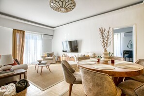 A modern living room with a dining area, a flat-screen TV mounted on the wall, and a large window with curtains. - SMG Boutique Apartment (Athina)