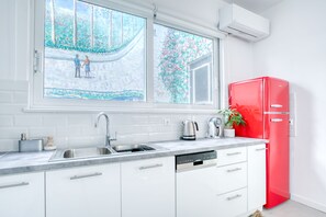 A modern kitchen with a stainless steel sink, white cabinets, a red refrigerator, and a window with a view of a garden. - SMG Boutique Apartment (Athina)