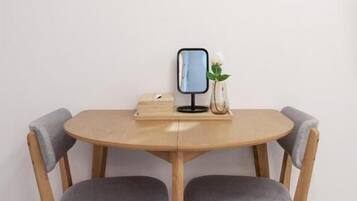 A round wooden dining table with two grey upholstered chairs, a framed artwork above, and a small vase with a plant on the table.