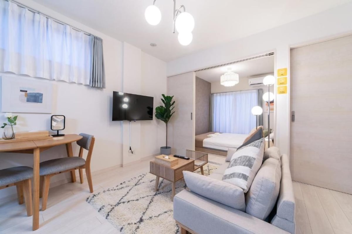 A modern hotel room with a flat-screen TV, a dining area with a wooden table and chairs, a sofa, and a large mirror.