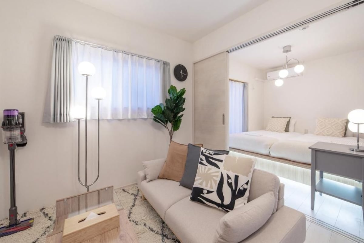 A modern living room with a sofa, a coffee machine, a plant, and a large mirror reflecting a bed and a desk area.