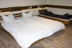 A double bed with a wooden headboard and a bedside table, a smaller single bed, and a wall-mounted shelf with a sign. - Cuckoo House Premium (Titisee-Neustadt)