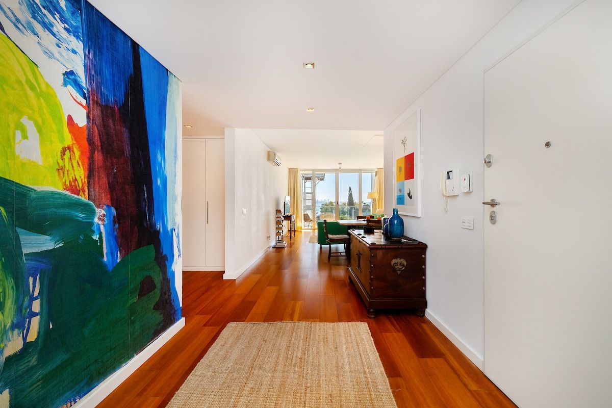 A hallway with a large abstract painting, wooden flooring, a rug, a desk with a vase, and a door with a light switch.